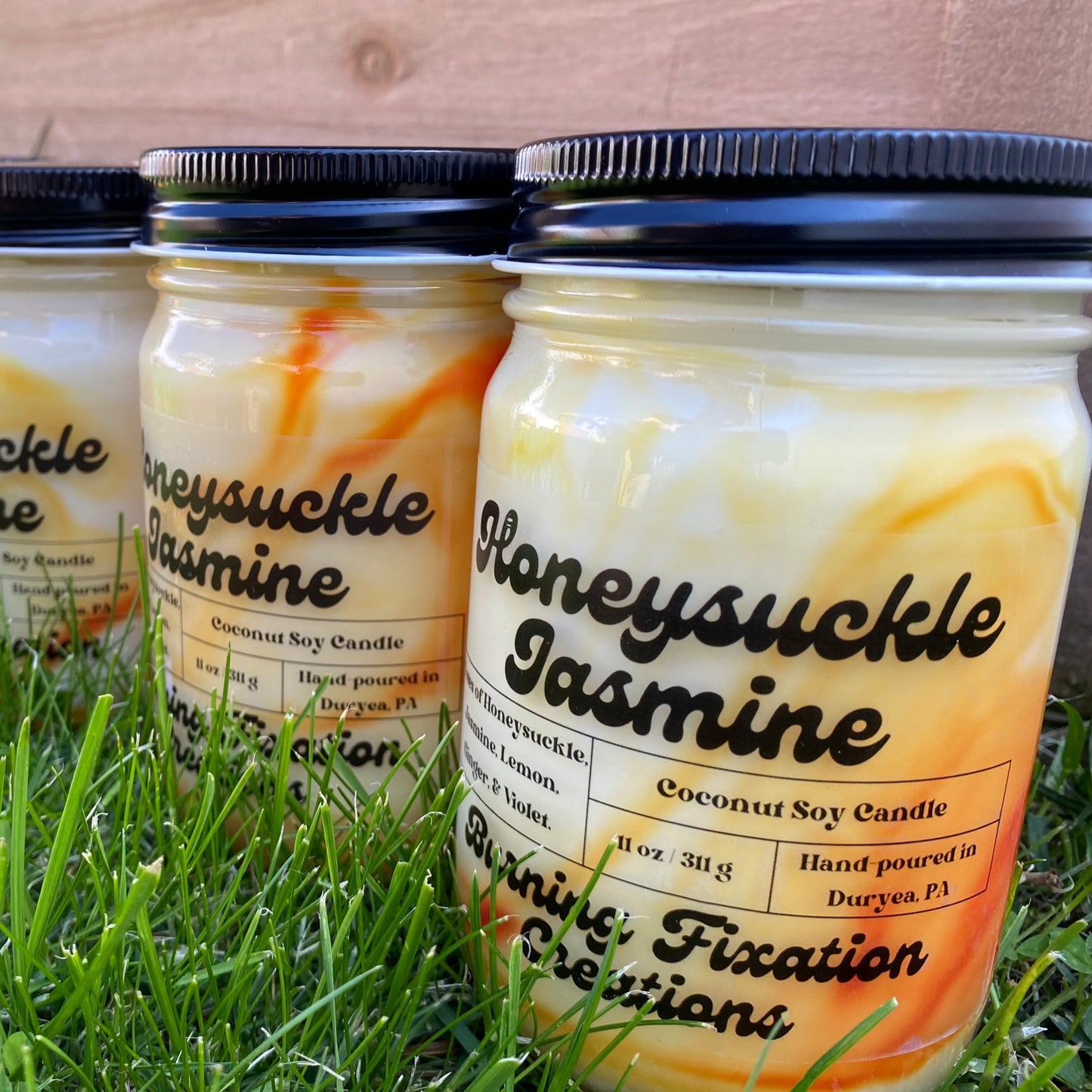 Honeysuckle Jasmine coconut soy scented candle with clear matte label, yellow and orange marbled in 12oz clear glass reusable jar with black button top screw on lid. Outdoor photo on green grass with wood fence in background.