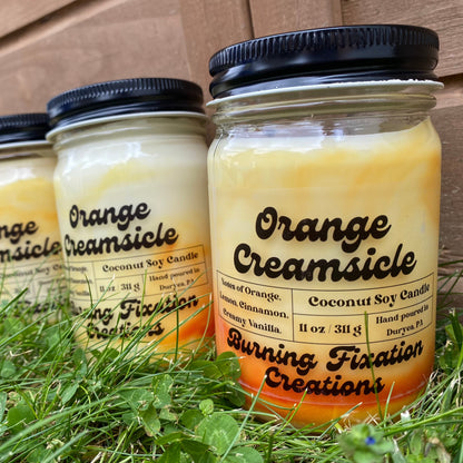 Orange Creamsicle coconut soy scented candle with clear matte label, orange and white marbled in 12oz clear glass reusable jar with black button top screw on lid. Outdoor photo on green grass with wood fence in background.