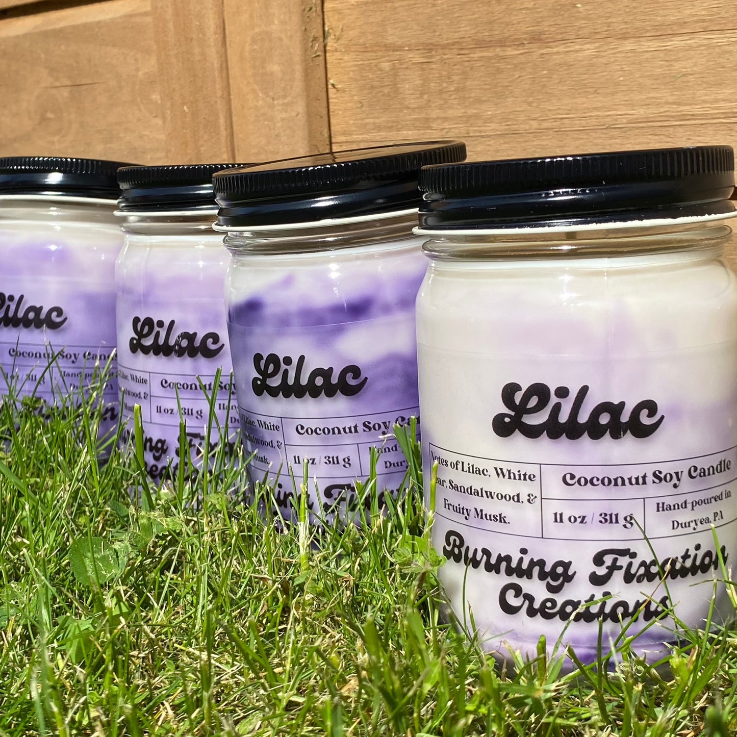 Lilac coconut soy scented candle with clear matte label, purple and white marbled in 12oz clear glass reusable jar with black button top screw on lid. Outdoor photo on green grass with wood fence in background.